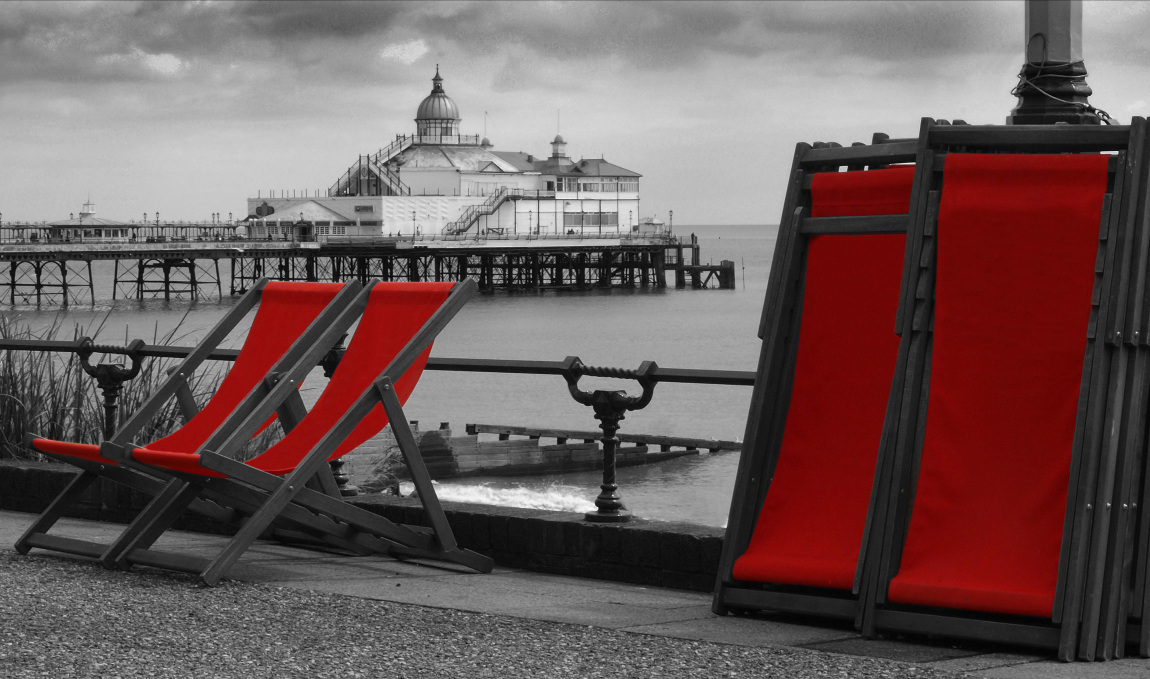 Enhanced black and white photograph of Eastbourne pier in the centre with a foreground of the promenade, two deckchairs ready for seating and several stacked deckchairs. All deckchair covers are picked out in blood red. © Copyright 2014 Andy Huntley photography